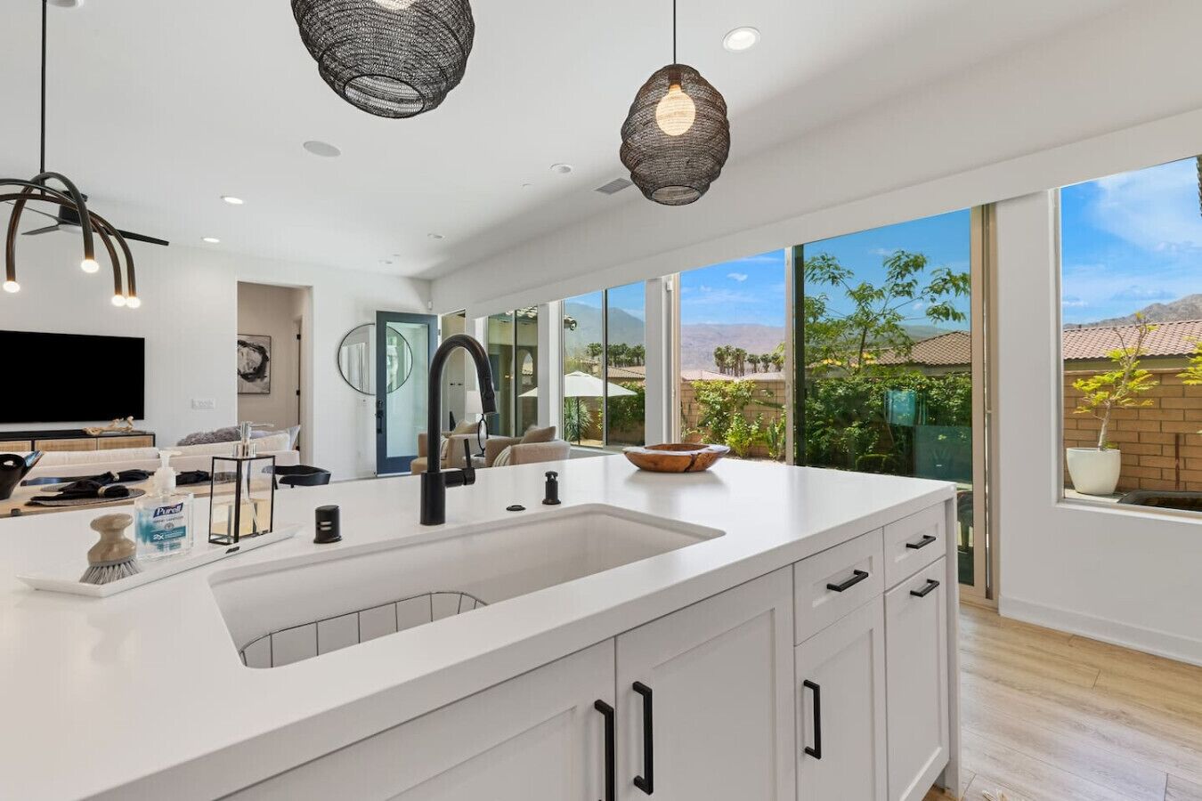 A modern kitchen with a large island, white cabinetry, and a view of a garden through large windows.