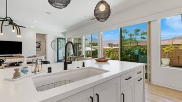A modern kitchen with a large island, white cabinetry, and a view of a garden through large windows.