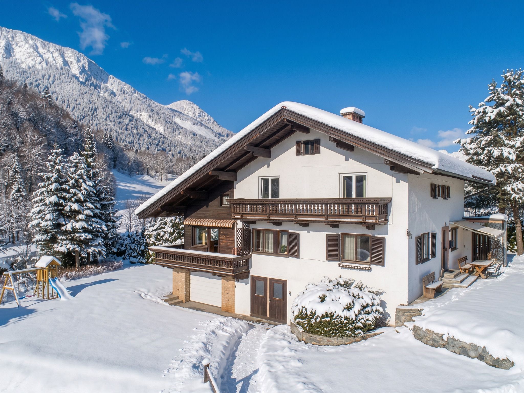 A two-story chalet with a balcony, set in a snowy mountainous area.