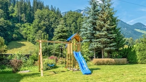 Zona verda amb un parc infantil, tobogan i cadiretes, i arbres, amb muntanyes al fons.