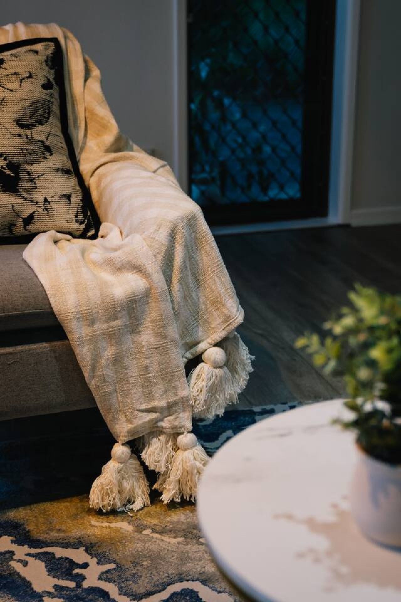 A sofa with a blanket and tassels, a patterned rug, and a potted plant.