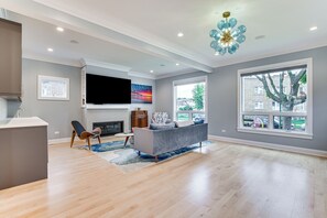 A spacious living room with hardwood floors, a grey sofa, a flat-screen TV, and a large window with a view of the outside.