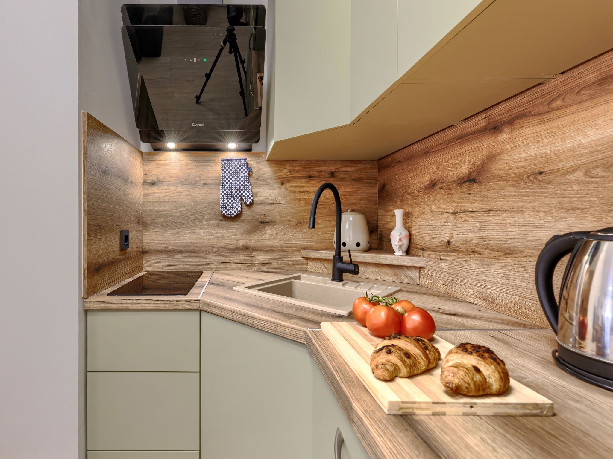 Una cocina moderna con protector de salpicaduras de madera, un lavabo empotrado, un hervidor y una tabla de cortar con tomates y croissants.