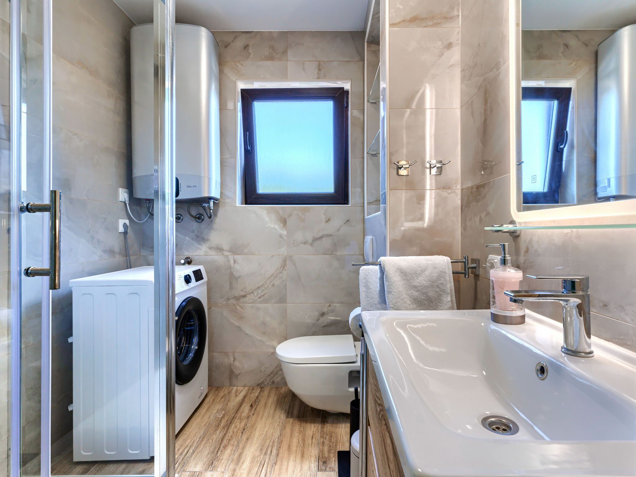 A modern bathroom with a built-in washing machine, a toilet, a sink with a mirror, and a window above the toilet.