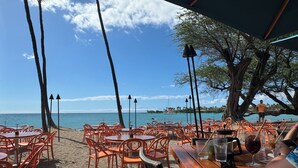 Een terras met oranje stoelen en tafels, met uitzicht op een strand waar een boot in de verte ligt.
