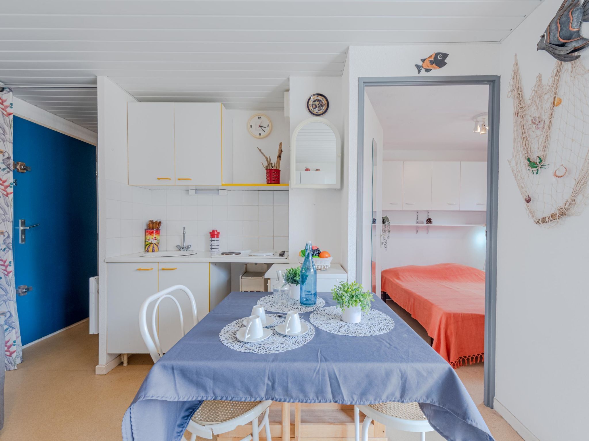 Une pièce compacte comprenant une table à manger pour quatre personnes, un coin cuisine, un lit recouvert d’une couverture orange et une petite fenêtre donnant sur la mer.