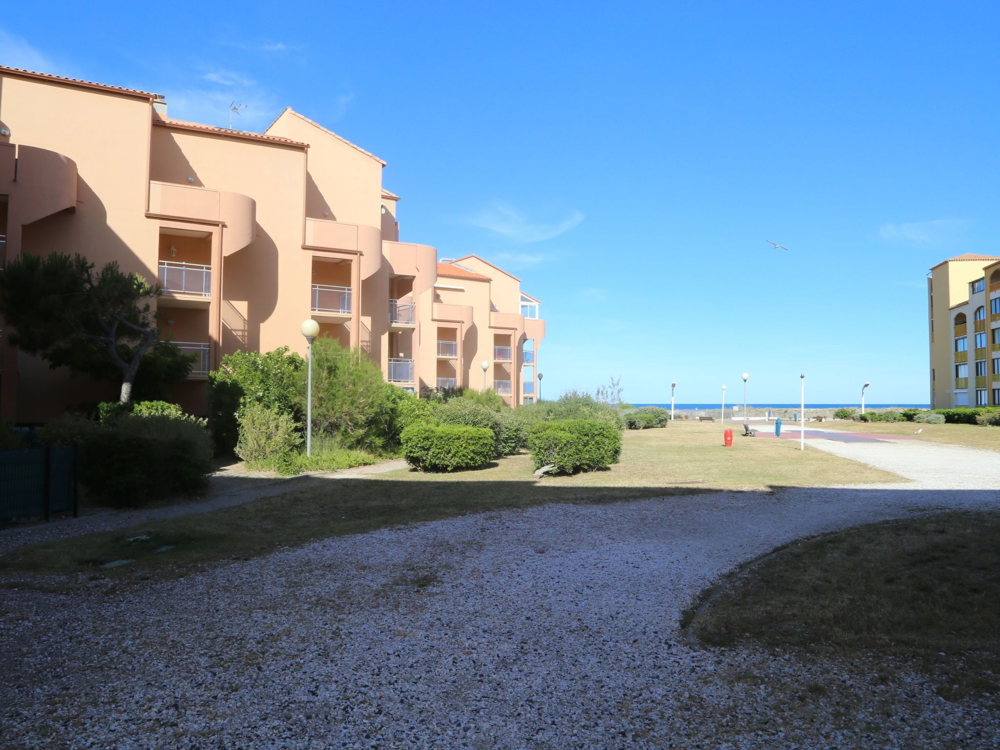 Un edificio de varios pisos con balcones, un patio de grava y una vista despejada del mar.