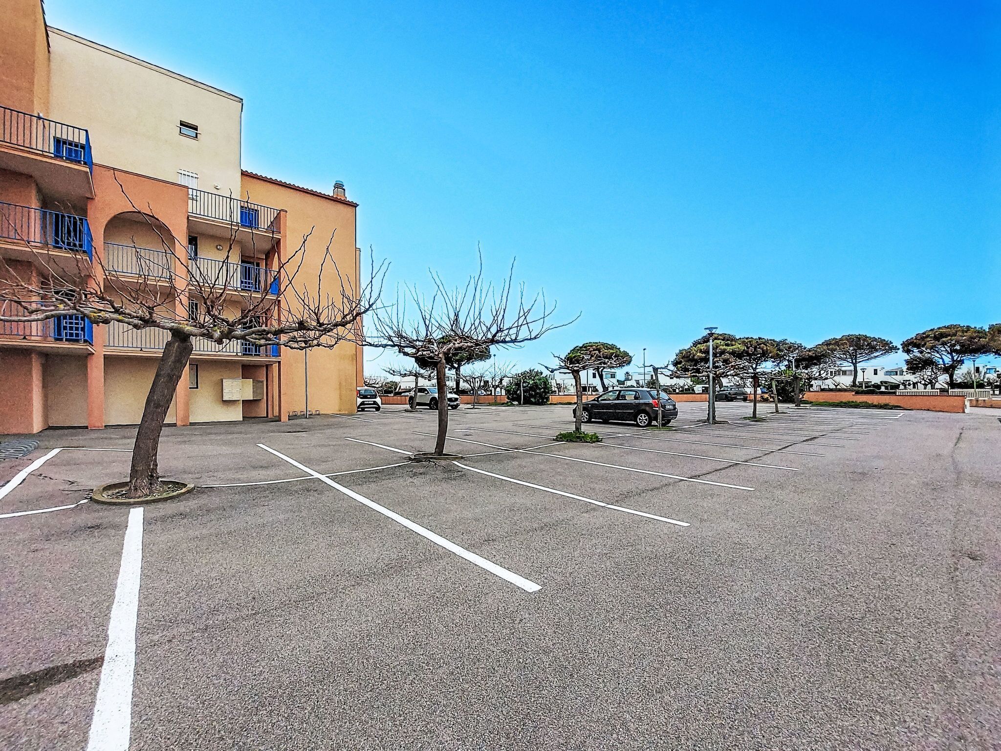 Un parking avec un bâtiment sur la gauche, quelques voitures et des arbres.