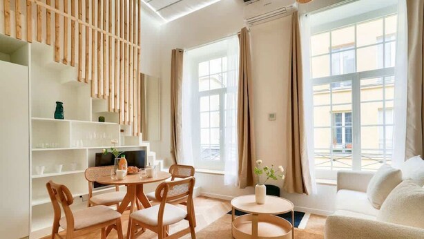 Una sala de estar moderna con una mesa redonda de comedor, sillas de madera, un sofá y una pequeña mesita auxiliar. Hay ventanas grandes con cortinas, una estantería y un estante montado en la pared con objetos decorativos.