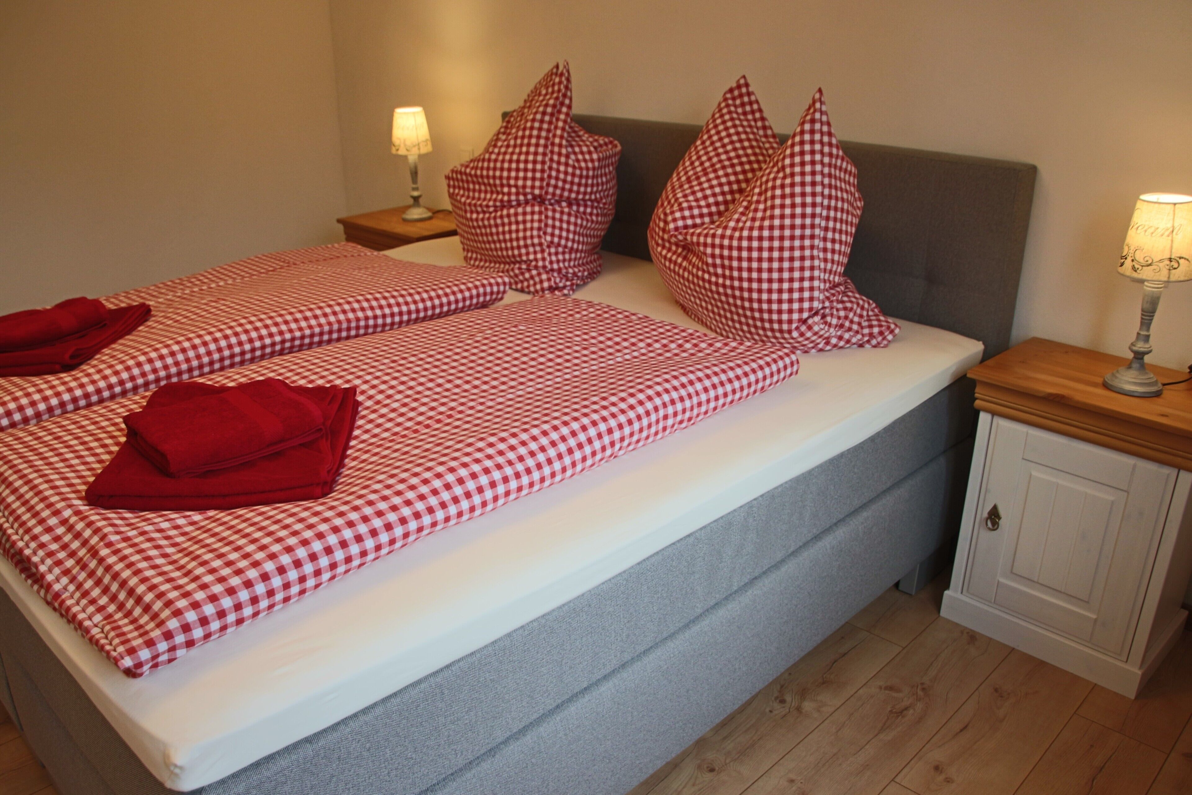Een bed met geruit beddengoed, kussens en een rode handdoek. Er staat een nachtkastje met een lamp en een kastje.