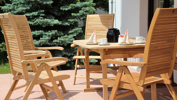 Conjunto de mesa e cadeiras de madeira ao ar livre, com vista para um arbusto verde.