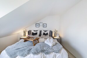 A bedroom with a sloped ceiling, a bed with a fluffy grey blanket, a bedside table with a lamp, and framed pictures on the wall.