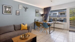A modern living room with a sofa, dining table, and chairs, a balcony with a view, and artwork on the walls.
