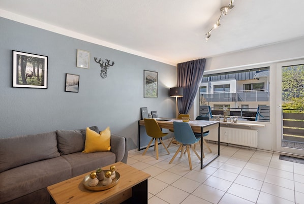 A modern living room with a sofa, dining table, and chairs, a balcony with a view, and artwork on the walls.