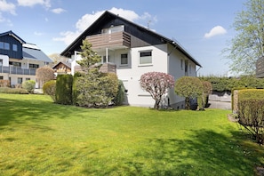 A modern two-story house with a balcony, surrounded by well-maintained lawns and shrubs.