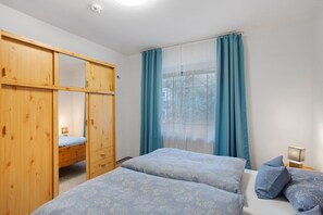 A bedroom with two beds, a wooden wardrobe, a window with blue curtains, and a bedside table with a lamp.
