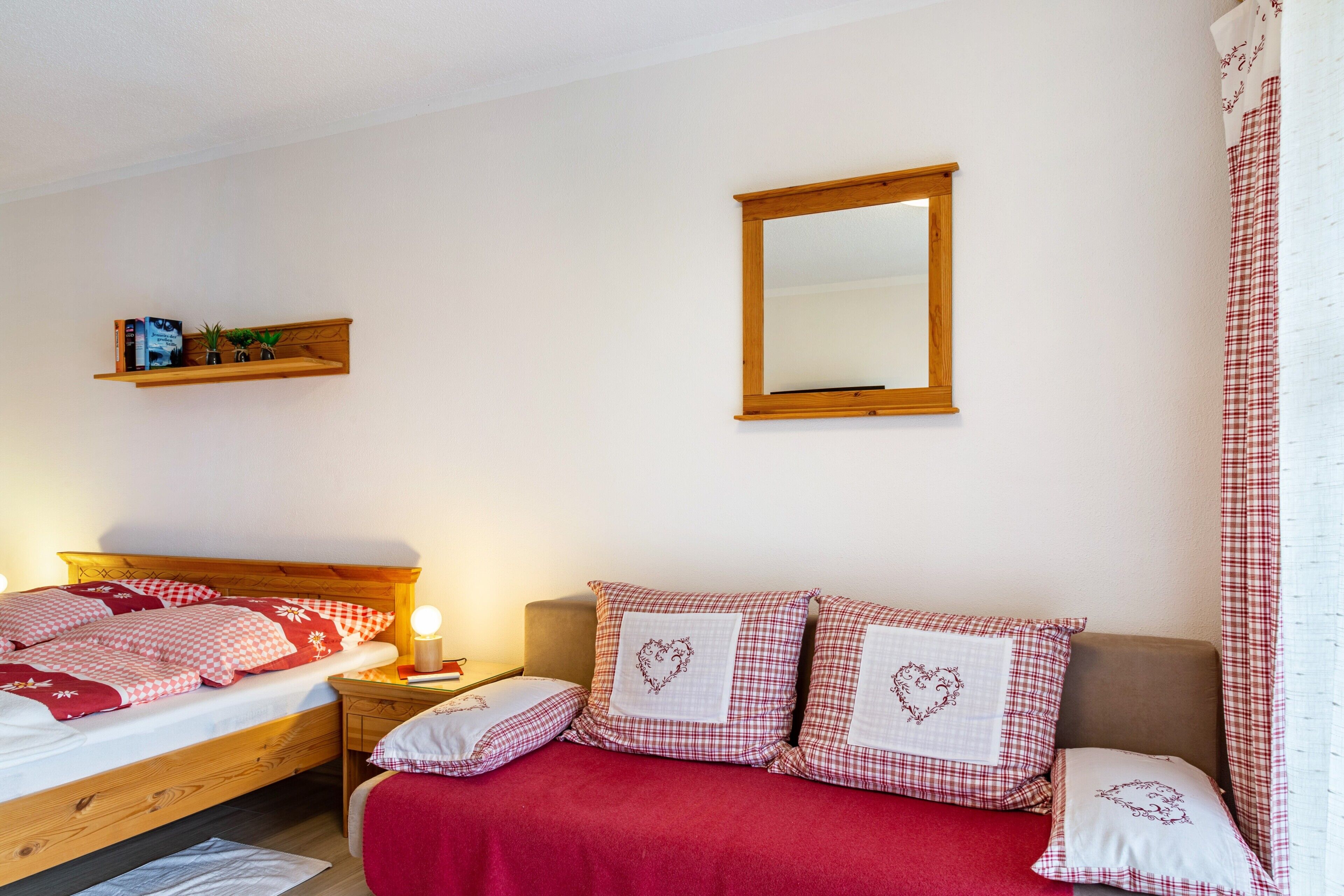 A bedroom with a wooden bed, a bedside table with a lamp, a mirror, and a shelf with books and plants.