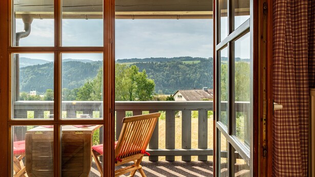 Un balcon avec une chaise et une table en bois, d’où l’on peut admirer un paysage boisé.