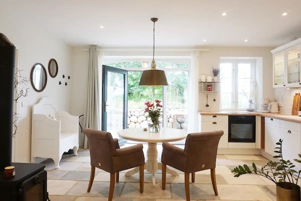 Un coin repas avec une table ronde et quatre chaises, une cuisine équipée d’un lave-vaisselle, et une vue sur la verdure à travers la porte vitrée.