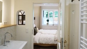 A bedroom with a bed, a window with a view, a bedside table, and a bathroom with a sink and mirror. - Apartment 'Lütje Orange' with Private Terrace, Shared Garden and Wi-Fi (Högel)