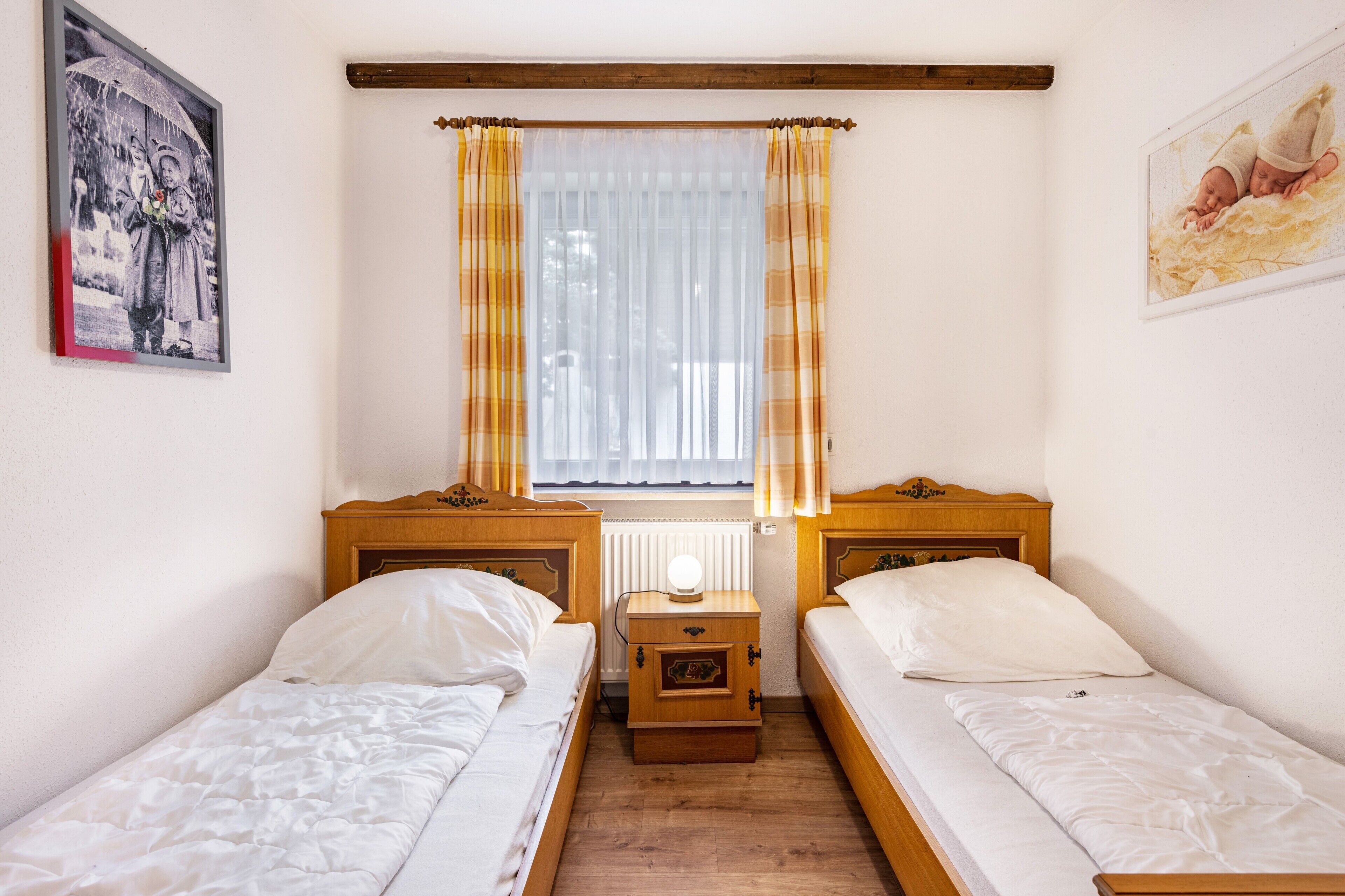 A room with two single beds, a nightstand with a lamp, a window with curtains, and wall art.