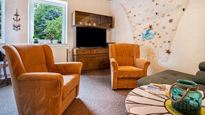 Un salón con dos sillones naranja, un televisor de pantalla plana, armarios de madera, un adorno de pared con conchas y vistas al verdor a través de un ventanal.