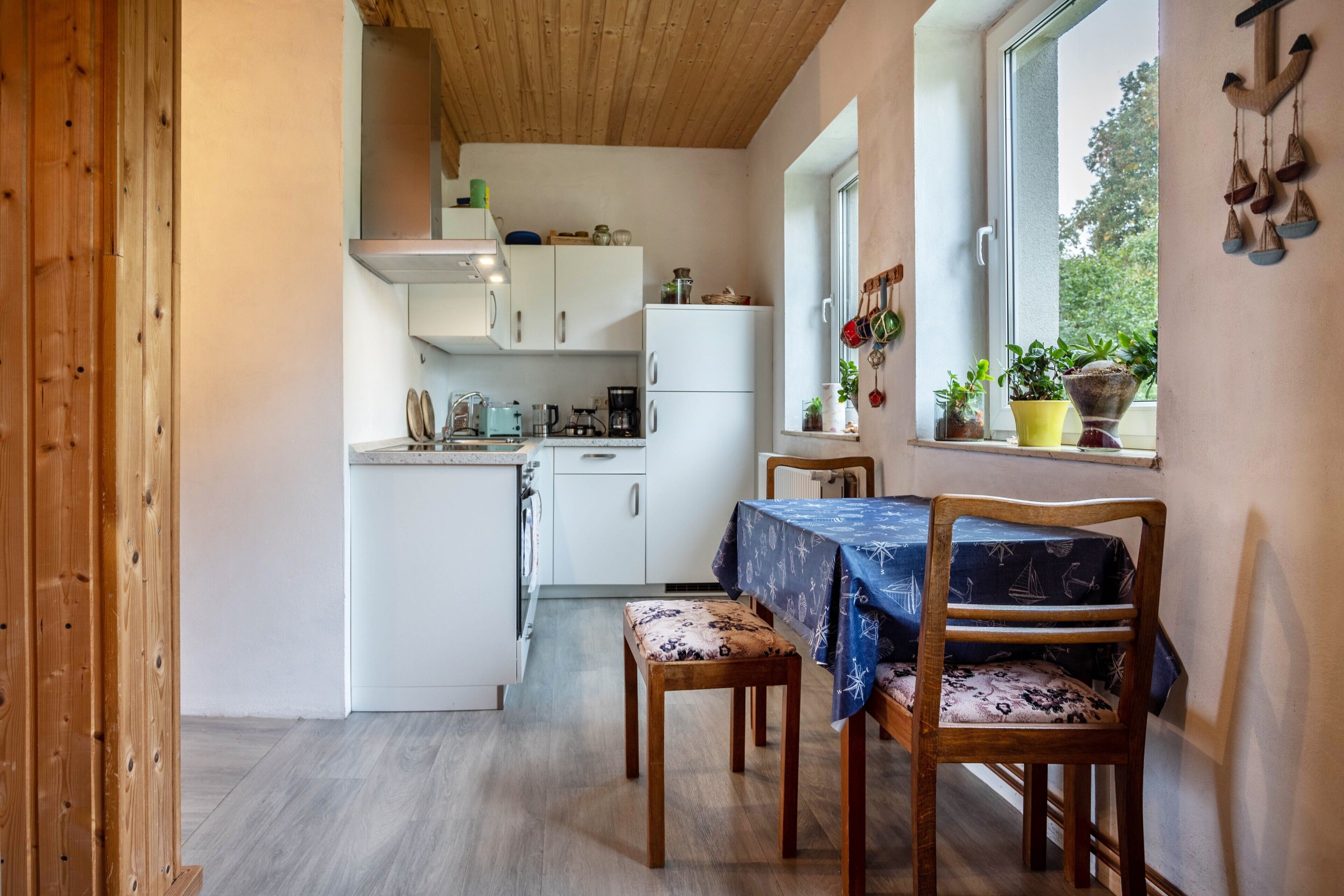 Une cuisine de taille réduite avec des meubles blancs, une table à manger avec des chaises et une fenêtre agrémentée de plantes.