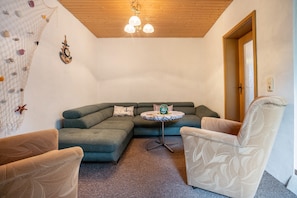 En stue med en grøn hjørnesofa, to møbelstole med mønster, et rundt bord med en dekorativ genstand, en klatrevæg og et træloft.
