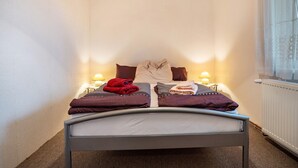 Una cama doble con dos almohadas, una manta y una toalla roja a la izquierda. Hay dos lámparas de noche a ambos lados de la cama.
