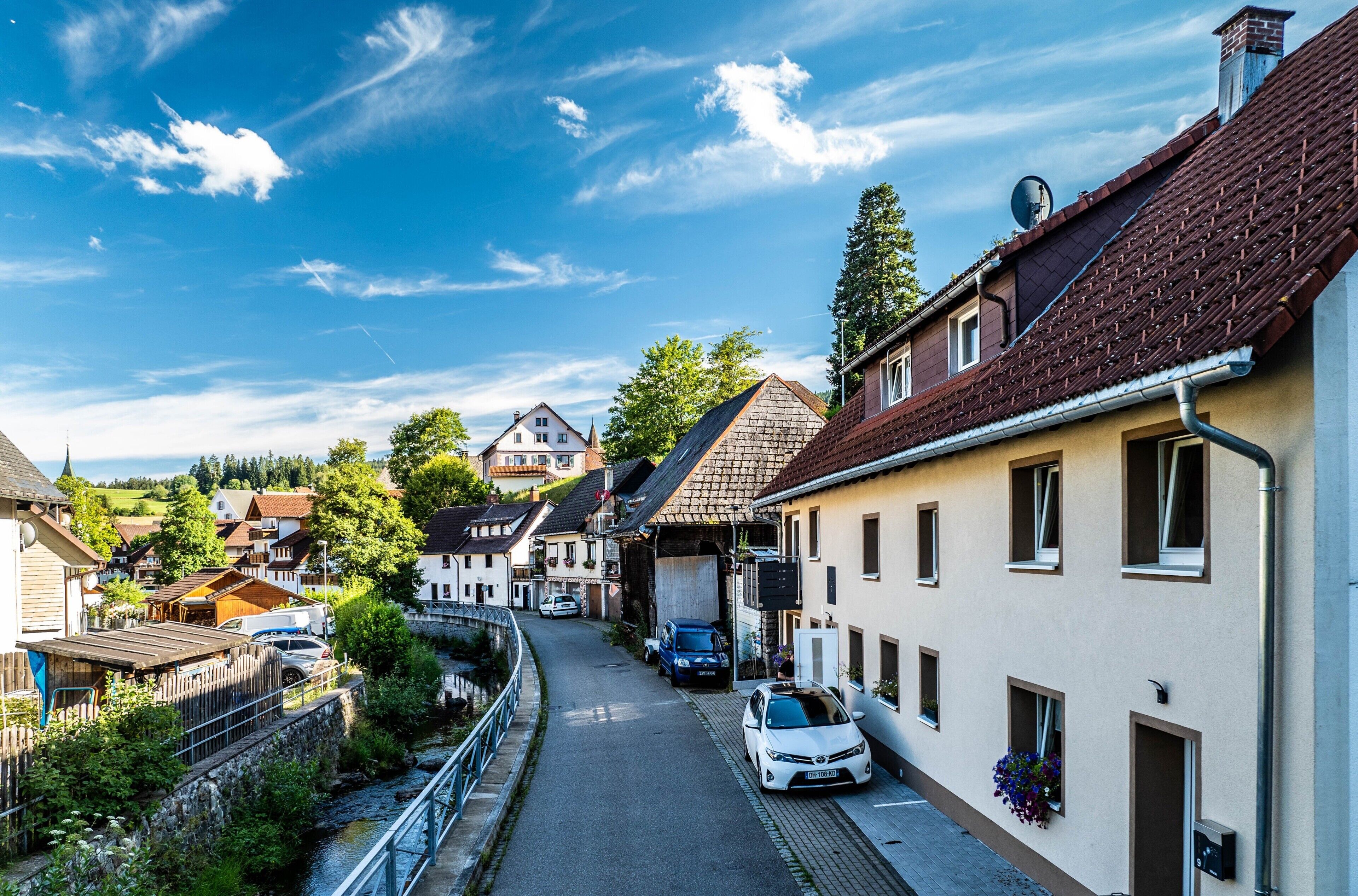 A narrow street lined with traditional buildings, a river on one side, and a clear blue sky.