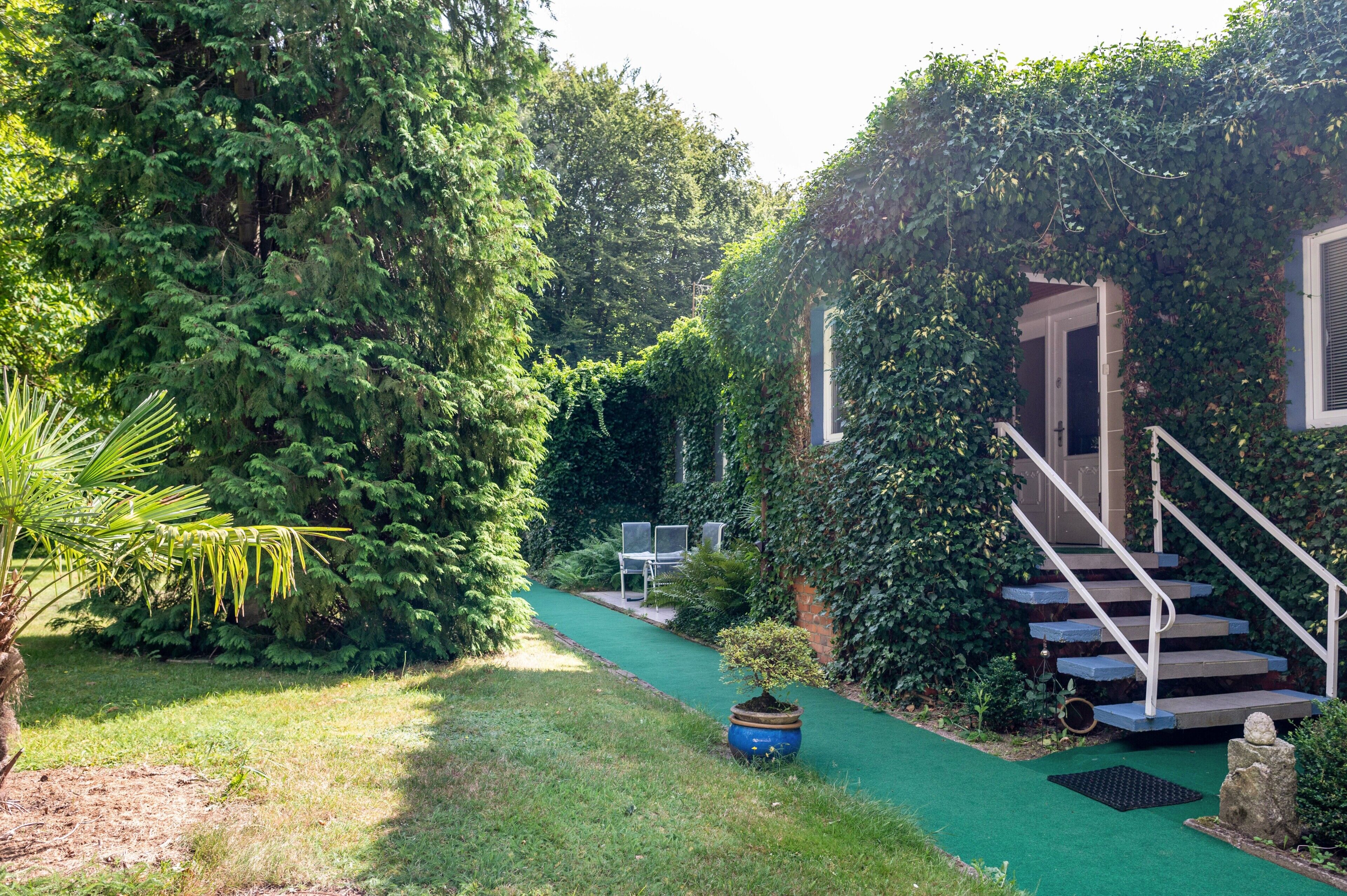 A garden pathway leading to a staircase with a metal railing, surrounded by lush greenery and a small tree in a pot.