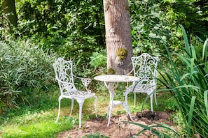 Un juego de mesa y sillas blancos en un jardín, con el tronco de un árbol al fondo.
