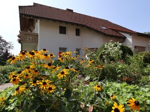 Un bâtiment doté d’un toit en tuiles, d’un balcon et d’un jardin où poussent des fleurs jaunes.