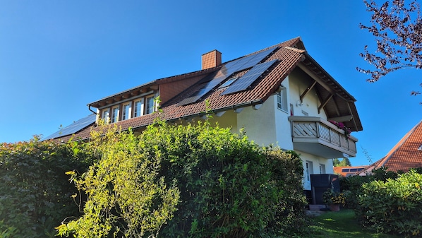 Une maison à deux étages avec un toit en pente, des panneaux solaires et un balcon orné de fleurs.