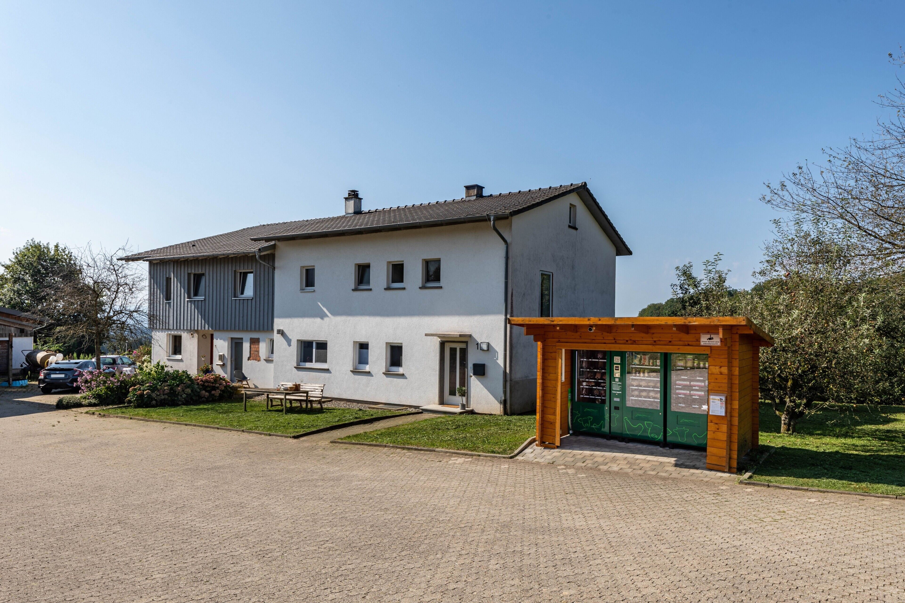 Un bâtiment à deux étages, doté d’une allée pavée, d’un petit espace extérieur aménagé pour s’asseoir et d’une structure en bois avec une porte verte.