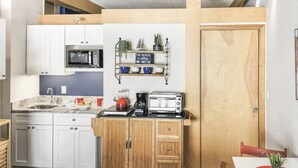 A compact kitchen with white cabinets, a microwave, a coffee maker, and a toaster. There is a wooden door and a small shelf with decorative items.