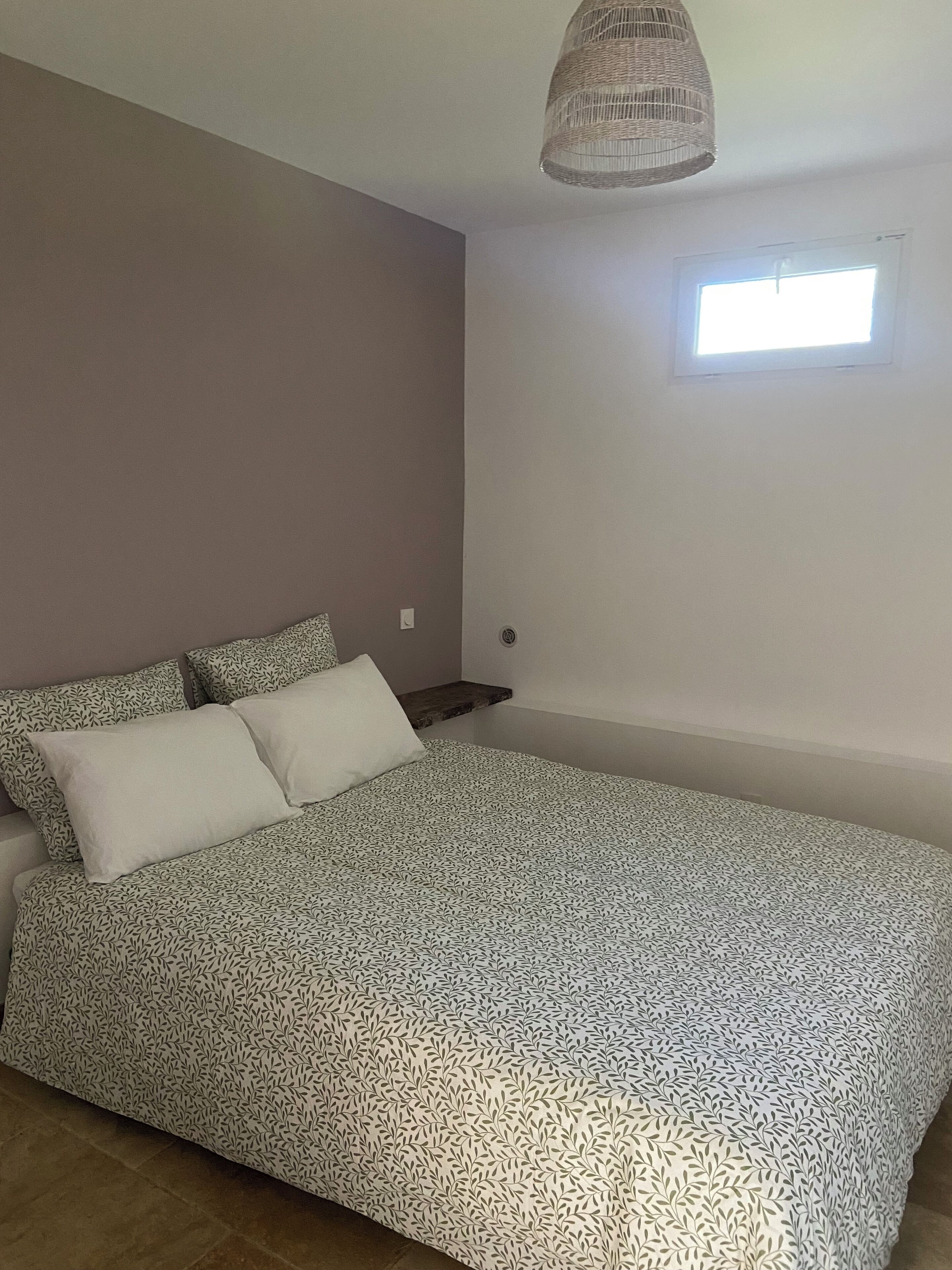 Een slaapkamer met een bed, kussens, een klein raam en een hanglamp.