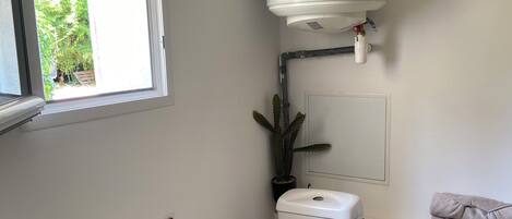 A small, clean bathroom with a toilet, a white water heater, a potted plant, and a window.