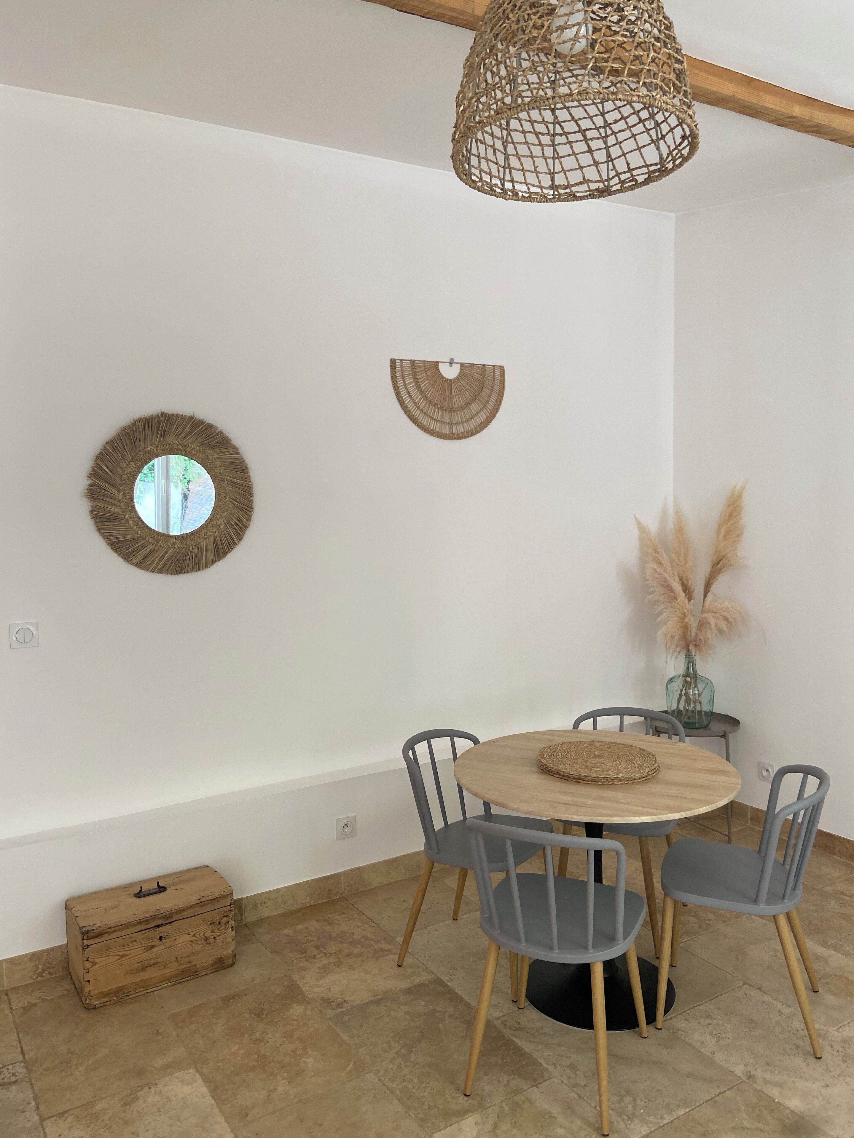 Un espace repas comprenant une table ronde en bois, des chaises grises, un luminaire suspendu en fibres tressées et deux miroirs fixés au mur.
