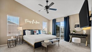 A bedroom with a bed, bedside tables, a desk, a chair, a ceiling fan, and a neon sign. - Desert Playhouse | Pool & Hot Tub • Arcade • Golf (Chandler)