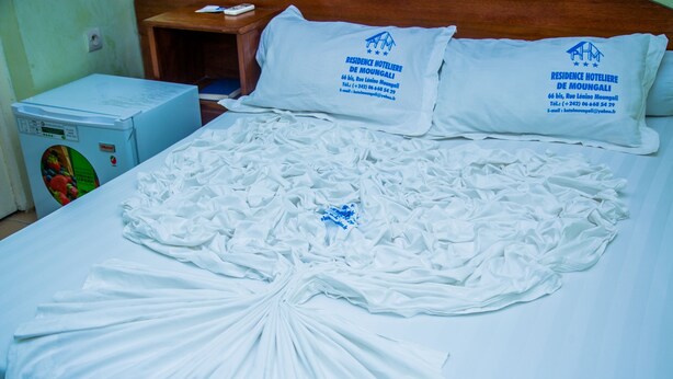 Una cama con sábanas y almohadas blancas, un refrigerador azul y una mesita de noche de madera con un control remoto.