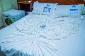 A bed with white sheets and pillows, a blue refrigerator, and a wooden nightstand with a remote control. - Résidence Hôtelière de Moungali (Brazzaville)