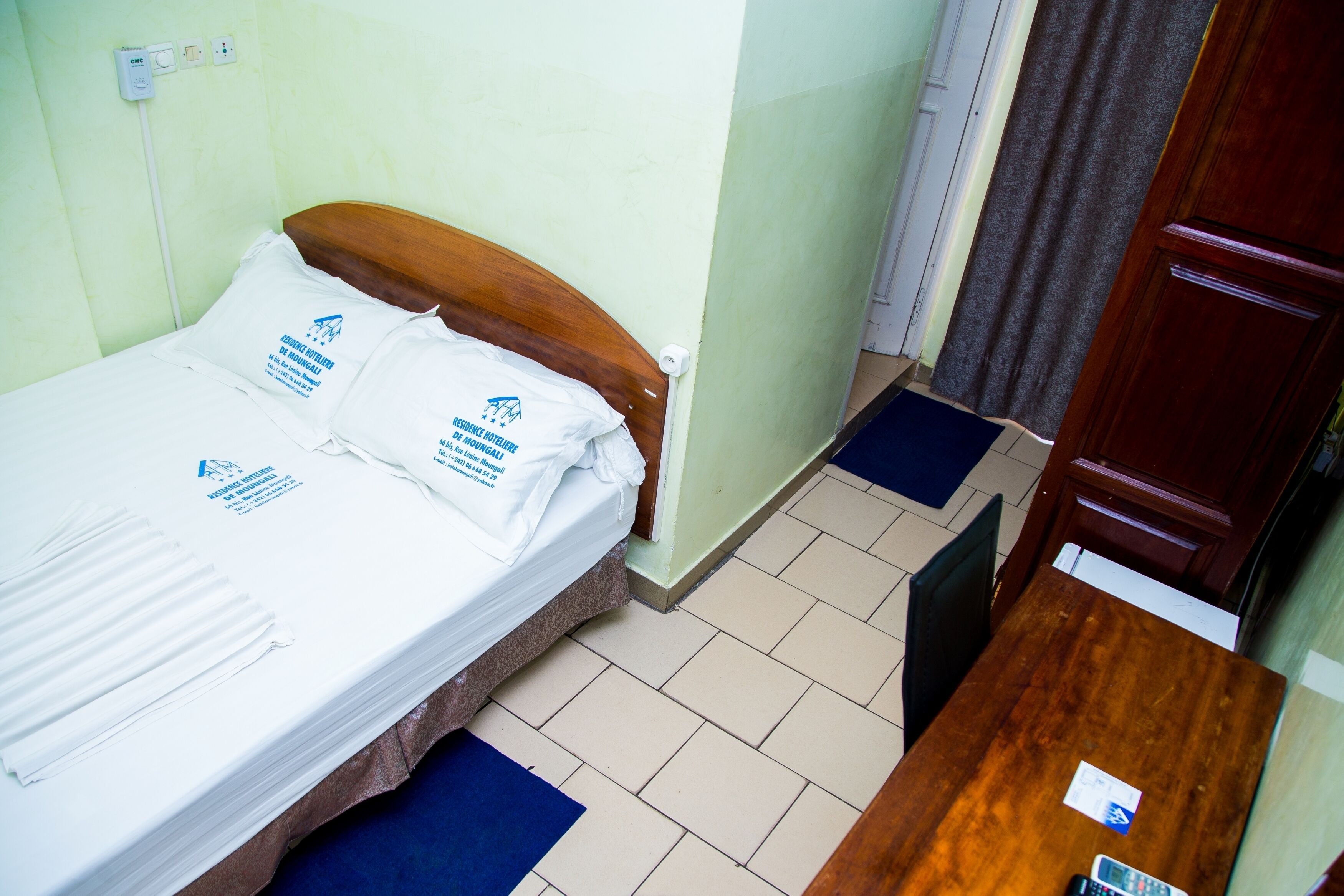 A single bed with white linens and pillows, a wooden headboard, a bedside table with a remote control, and a blue mat on the floor.