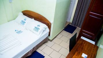 A single bed with white linens and pillows, a wooden headboard, a bedside table with a remote control, and a blue mat on the floor.