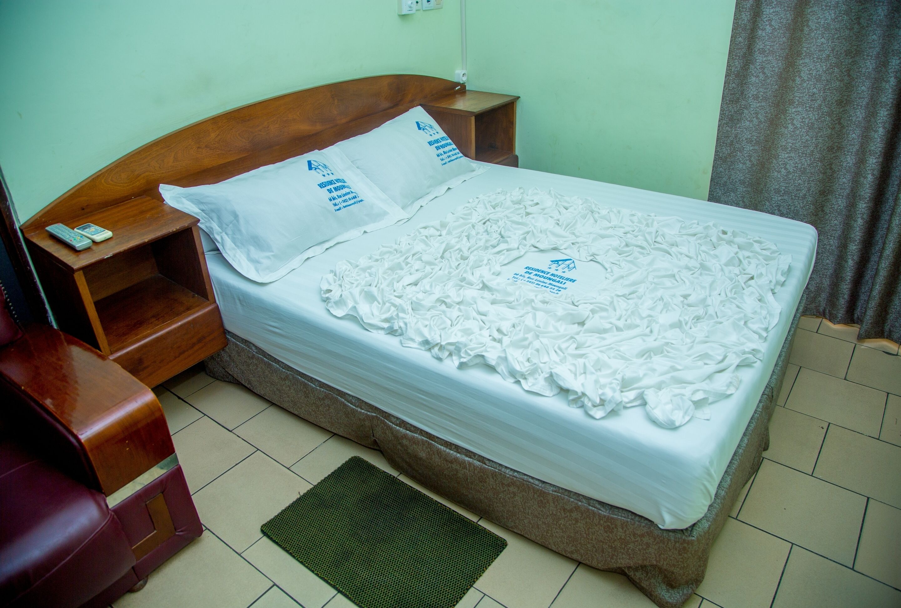 A neatly made bed with white linens and decorative pillows, a wooden nightstand with a phone, and a green mat on the floor.