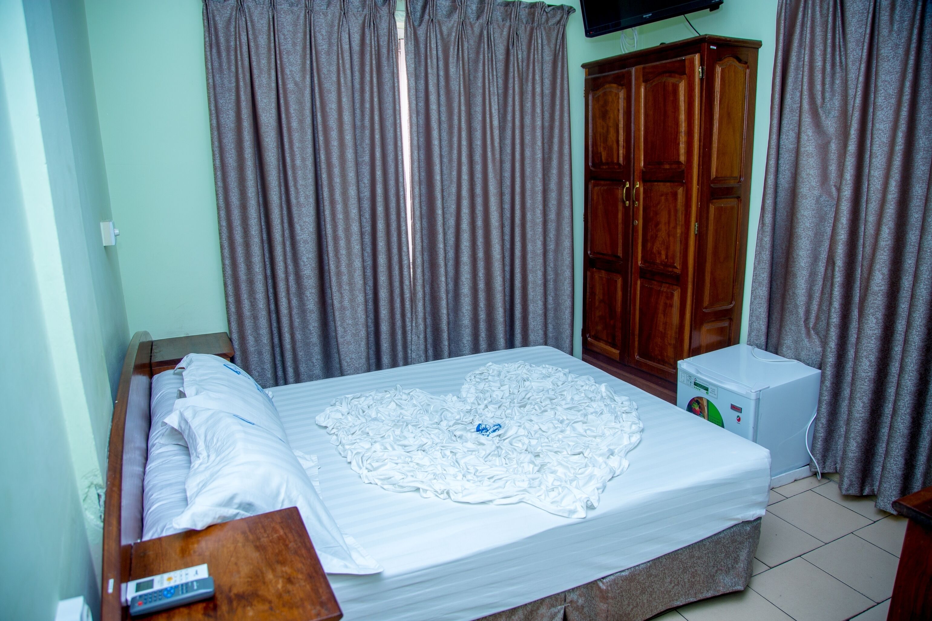 Una cama bien hecha con edredón blanco y almohadas, un armario de madera, un electrodoméstico pequeño blanco y cortinas grises.
