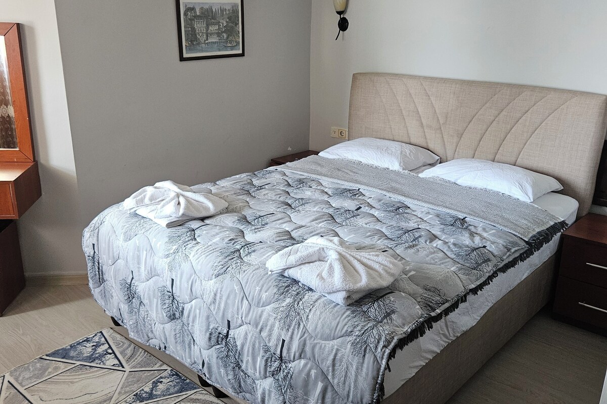 A neatly made bed with a patterned duvet, a bedside table, a wall-mounted lamp, and a framed picture on the wall.