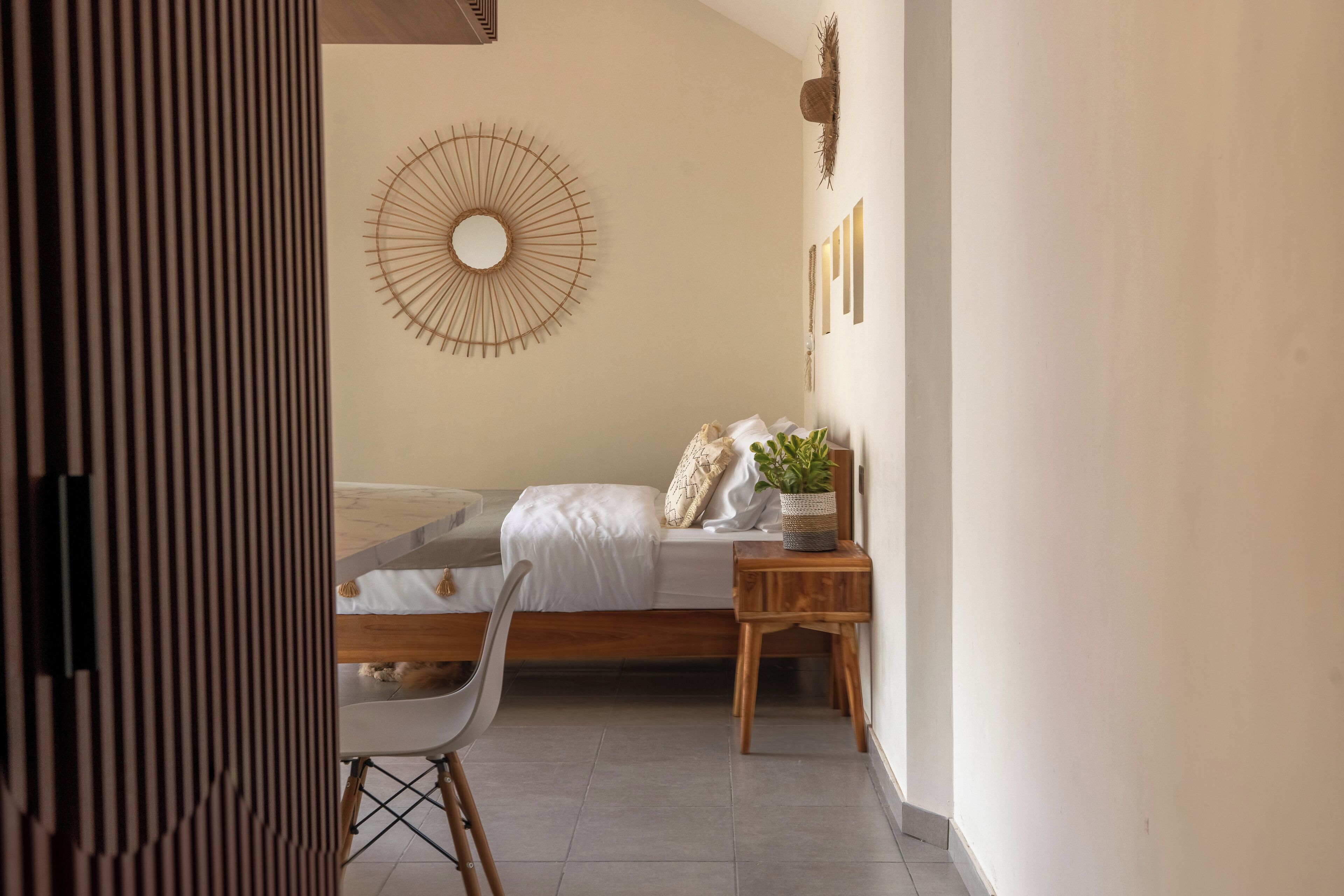 A bedroom with a bed, a bedside table with a plant, a chair, and a wall-mounted circular mirror.