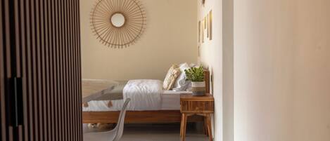 A bedroom with a bed, a bedside table with a plant, a chair, and a wall-mounted circular mirror.