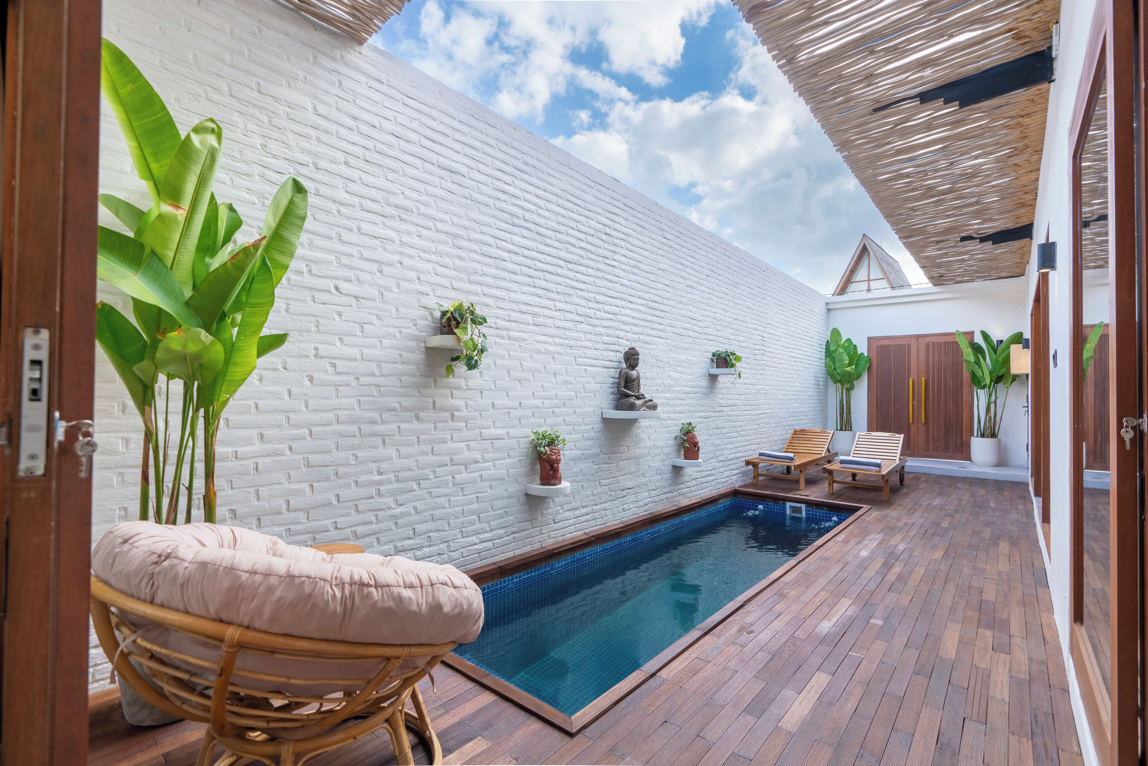 Un área de piscina con pared de ladrillo blanco, terraza de madera y sillones de descanso.